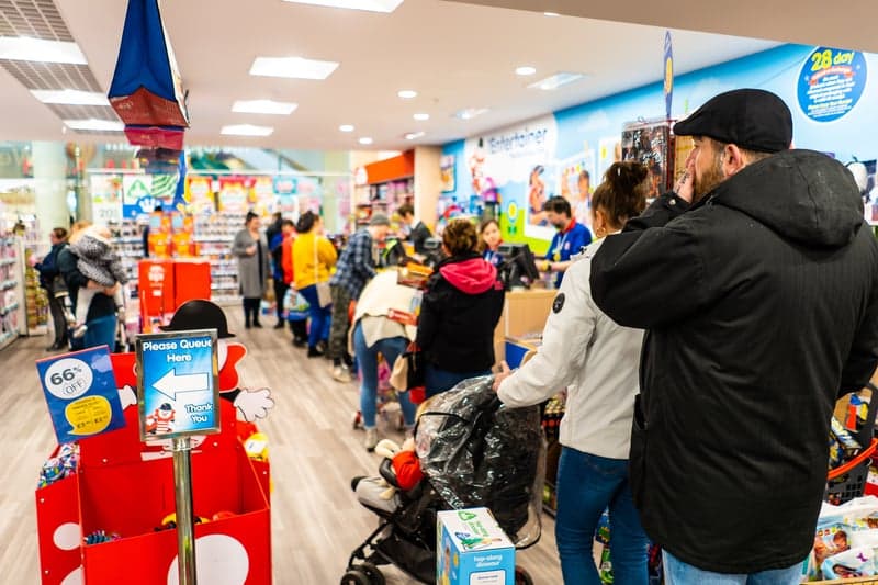 Warteschlange in einem Geschäft, davor ein Schild mit der Aufschrift „Please Queue Here“